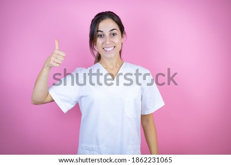 Young brunette doctor girl wearing nurse or surgeon uniform over isolated pink background smiling and doing the ok signal with her thumb