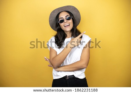 Beautiful woman wearing sunglasses, casual white t-shirt and a hat standing over yellow background smiling pointing to the side