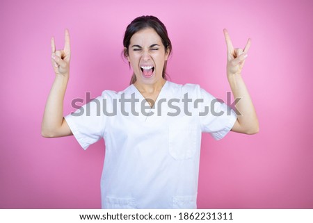 Young brunette doctor girl wearing nurse or surgeon uniform over isolated pink background shouting with crazy expression doing rock symbol with hands up