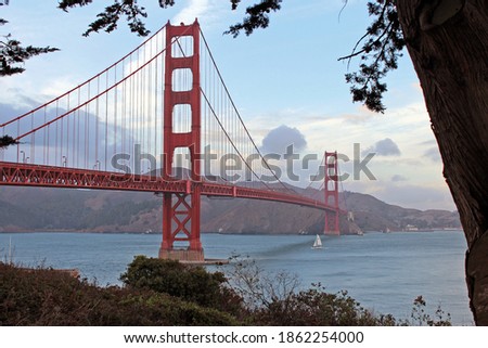 Golden gate bridge on a sunny summer day. Picture taken through an opening between some trees. Boat passing underneath. Blue skies with some clouds.