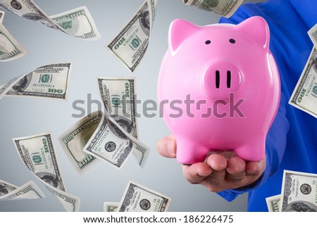 Businessman hand holding pink piggy bank and one hundred dollar banknotes flying on dark background.