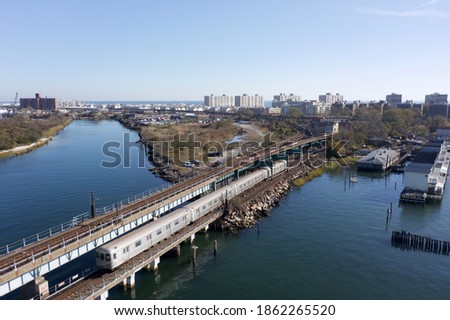 An aerial shot above train tracks in Queens, NY. There is a train on the tracks crossing over the bay. It is a sunny afternoon and the water is calm and reflective.