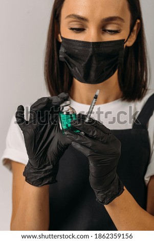 a girl in a black mask and black gloves is holding a bottle with a green drug and a syringe. photo in studio on white background