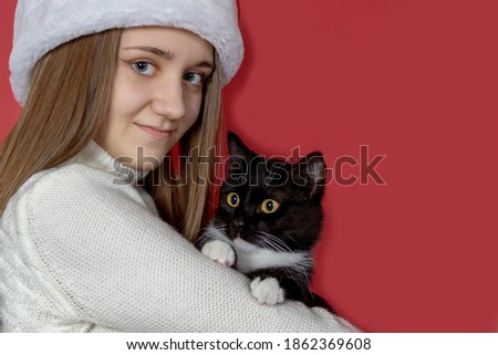 A beautiful smiling girl in a red Santa hat and a black cat in her hands on a red isolated background, close-up, copy of the space, selective focus. New year or Christmas celebrations