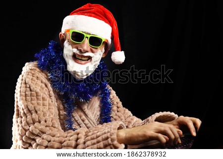 Bad crazy Santa in sunglasses, foam beard in home clothes sitting at a laptop