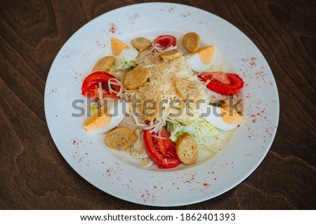 Salad with croutons, tomatoes and eggs, sprinkled with cheese and cabbage. Snack on a white round plate on a dark wooden background