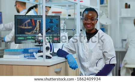 Professional black woman scientist looking at camera smiling in modern equipped lab. Multiethnic team examining virus evolution using high tech and tools for scientific research, vaccine development. Royalty-Free Stock Photo #1862409934