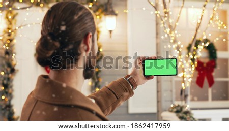 Close up shot of black smartphone with green screen in hand while standing outdoors on Christmas Eve. Mobile phone with chroma key in man fingers in winter. Technology concept