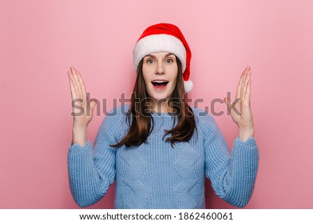 Shocked cute woman in Christmas hat shows something large and of big size, measures with hands and keeps mouth opened from wonder, talks about something huge, wears sweater, models over pink wall