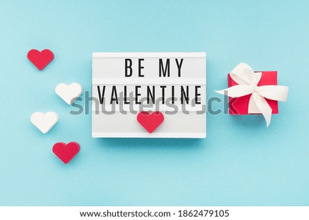 Valentine's Day holiday card. Red gift box, hearts on pastel blue background. Valentines day concept. Flat lay, top view, copy space. Text Be My Valentine on white lightbox.