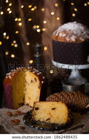 Two Italian Christmas bread Panettone, with one sliced and placed on a desk with fairy lights at the background and pine cone on the desk, Christmas atmosphere