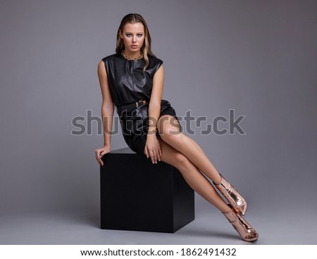 Fashion photo of a beautiful elegant young woman in a pretty leather dress, bronze-peach metallic boots, thick metal chain around her neck posing over gray background. Studio shot, portrait. 