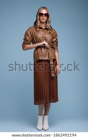 Fashion photo of a beautiful elegant young woman in a pretty brown leather jacket, brown long skirt, stylish sunglasses  posing over blue background. Studio shot, portrait. Brown and blue colors