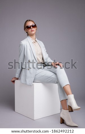 Fashion photo of a beautiful elegant young woman in pretty soft blue suit, jacket, pants, trousers, white boots, stylish sunglasses posing on gray background. Studio shot, portrait. Monochrome colors