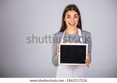 Female Employee Wearing Uniform Apron Holding Blackboard Sign. Friendly smiling shop assistant holding blank chalkboard sign isolated on gray background. 