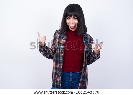 Born to rock this world. Joyful Young Caucasian beautiful woman wearing plaid shirt against white background screaming out loud and showing with raised arms horns or rock gesture.