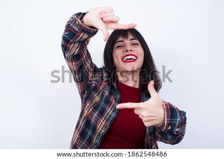 Positive Young Caucasian beautiful woman wearing plaid shirt against white background with cheerful expression, has good mood, gestures finger frame actively at camera.