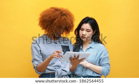 Young multiracial friends shopping online using tablet. Afican american and asian women isolated on yellow background. High quality photo