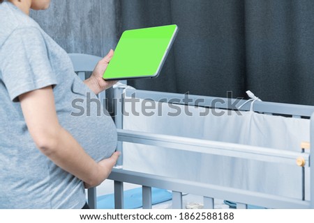 A pregnant girl stands near a crib and uses a digital tablet with a green screen for chromakey consults online, empty space