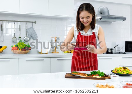 Asian woman chef in red apron take a picture of salmon steak with smartphone. Concept woman preparing meals at home.