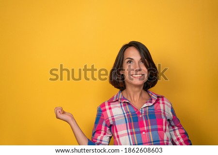 OK or Okay sign with one raised hand smile beautiful woman dressed in plaid shirt and dark hair on a yellow background. Human emotions, facial expression concept.