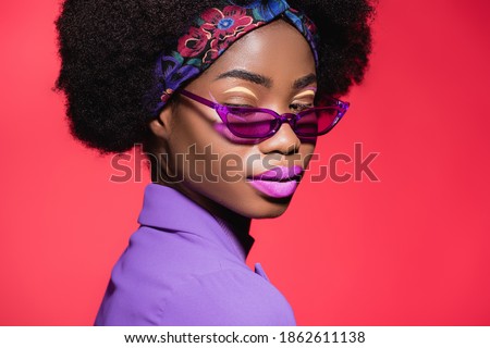 african american young woman in purple stylish outfit and sunglasses isolated on red