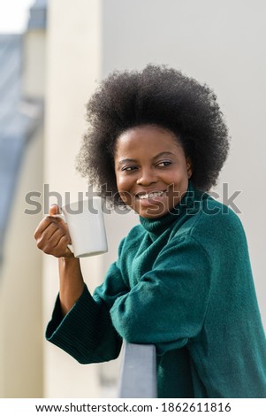 Smiling young African American biracial woman enjoying a cup of tea or coffee, wear oversize green knitted sweater, looking away, standing on the balcony. 