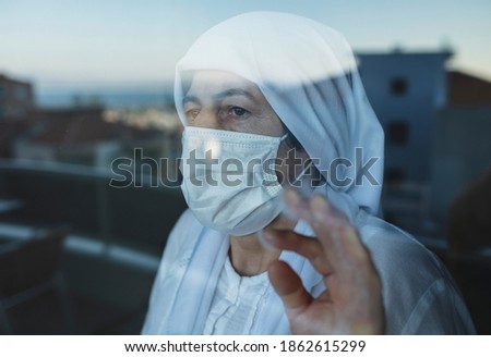 An elderly woman in quarantine at Christmas time wears a surgical mask and looks out the window.