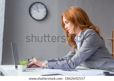 Side view of focused female lawyer typing on laptop, while sitting at workplace on blurred background