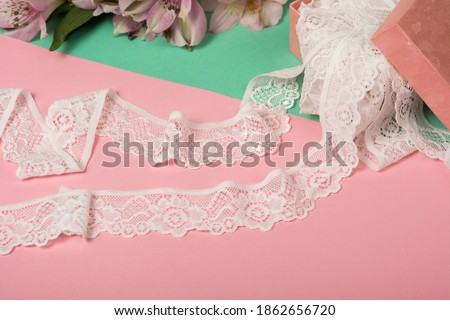Delicate tapes of white gentle ribbon, beauty lace fabric with furniture and flowers, accessories, bands, rings on pink and green background. Elastic material. Using for Atelier and needlework store.