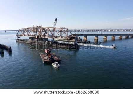 A low angle drone shot of a swing bridge that is opened to allow a tall dredge to pass. It is a sunny say in the afternoon and the skies are blue. The water of the bay is calm.