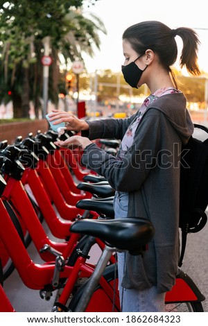 vertical photo of a young girl with protective mask cleaning her hands with disinfectant gel next to an urban bicycle rental parking, concept of protection against covid and sustainable mobility