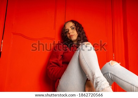 Young woman sitting on floor. Curly brunette poses near red wall.