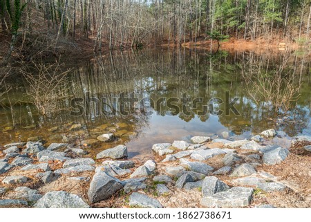 A small lake with reflections of the woodlands on the water on a bright sunny day with a sitting bench and birdhouse in the background and large boulders in the foreground