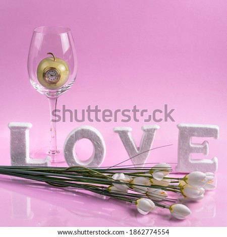 The creative romantic concept with letters. A glass with an Apple clock inside and flowers in front of letters. Pink background.