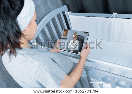 A pregnant girl stands near a baby's crib and uses a digital tablet to consult a doctor online. The concept of social distancing during the Covid 19 quarantine