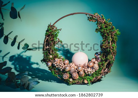 Intricate round, modern Christmas and winter season arrangement with a candle, made out of twigs, moss, glass bals, and dry pine cones on a matte background.  