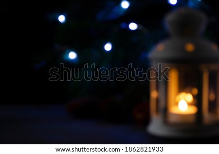 Blurred picture for background. White lantern with a burning candle, pine branch and garland lights on background. Christmas mystery mood.