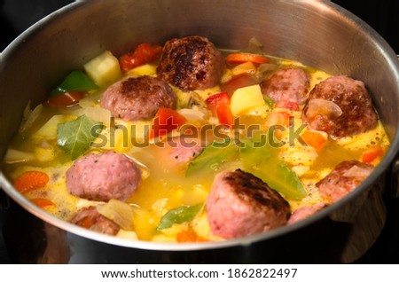 Pot with stew, meatballs, potatoes, carrots, green peppers and bay leaves being cooked on a ceramic stove.