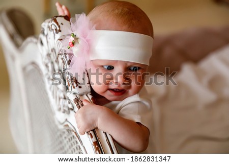 Cute happy blue-eyed 6-month-old girl in white headband at head of retro bed. Pretty smiling blonde beauty. Charming child shows emotions in close-up. Concept of a healthy child and parenting