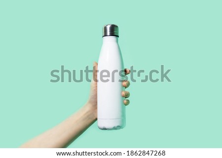 Close-up of female hand, holding white reusable steel stainless eco thermo water bottle on background of cyan, aqua menthe color. Be plastic free. Zero waste. Royalty-Free Stock Photo #1862847268