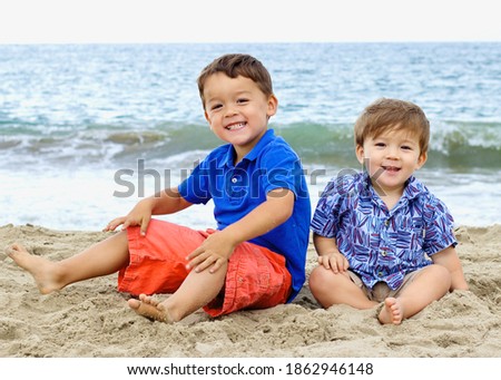 Two Japanese American brothers sitting on a beach with the ocean background. The older brother is wearing a blue polo shirt and orange shorts. The younger one is wearing a blue button up and khakis. 