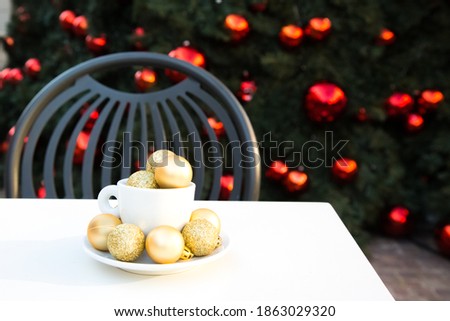 Composition with coffee cup, golden Christmas balls inside and around. Coffee pair on white table angle, grey chair, new year tree with red toys in background. Winter cafe. Post card picture. 