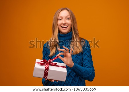 Portrait of a happy young woman in knitted blue sweater holding gift decorated with ribbon. Studio shot, yellow background. New Year, Women's Day, Birthday, Holiday concept