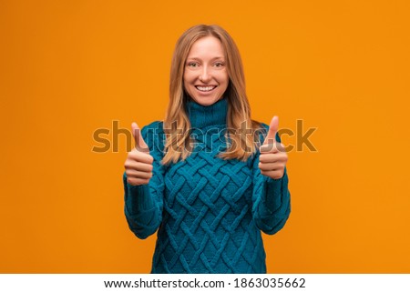 Young positive woman in knitted blue sweater smiling and showing a thumbs up gesture. Good job, happiness, body language, facial expression concept. Studio shot, yellow background, isolated