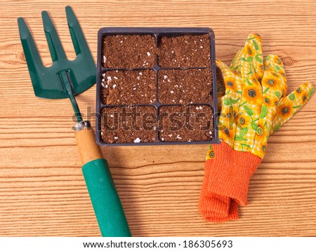 A pair of floral gardening gloves and a trowel and a six cell pack of seeds against a wooden background.