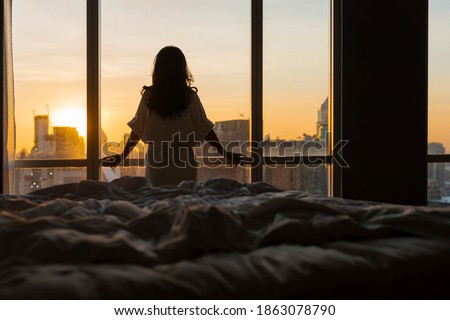 Beautiful asian woman is waking up in the morning, Sun shines on her from the big window. Happy young girl greets new day with warm sunlight flare and city scenery in the window. Co Royalty-Free Stock Photo #1863078790