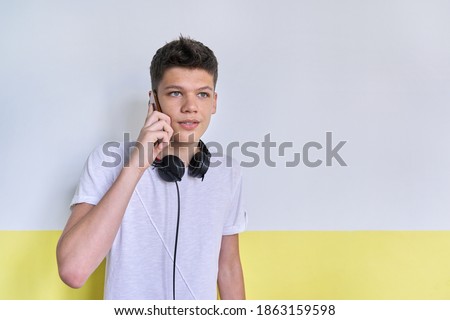 Portrait of student teenager 16, 17 years old in white t-shirt with headphones, talking on mobile phone, looking to the side, white wall background copy space