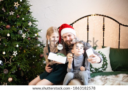 Bearded father with Santa Claus hat and his two children congratulate online their family using smartphone  or making selfie together holding gift box. Christmas online congratulations during pandemic