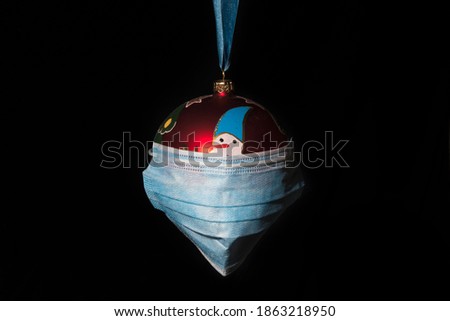 Symbolic picture for christmas time during covid pandemic (corona virus), a red christmas decoration ball with a covid19 face protection mask, haning on a blue fabric tape on black background